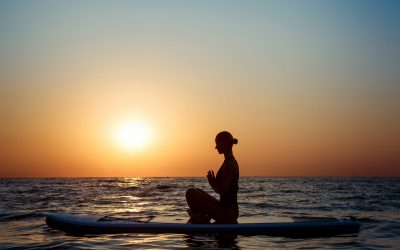 Silhouette of young beautiful girl practicing yoga on surfboard in sea at sunrise.